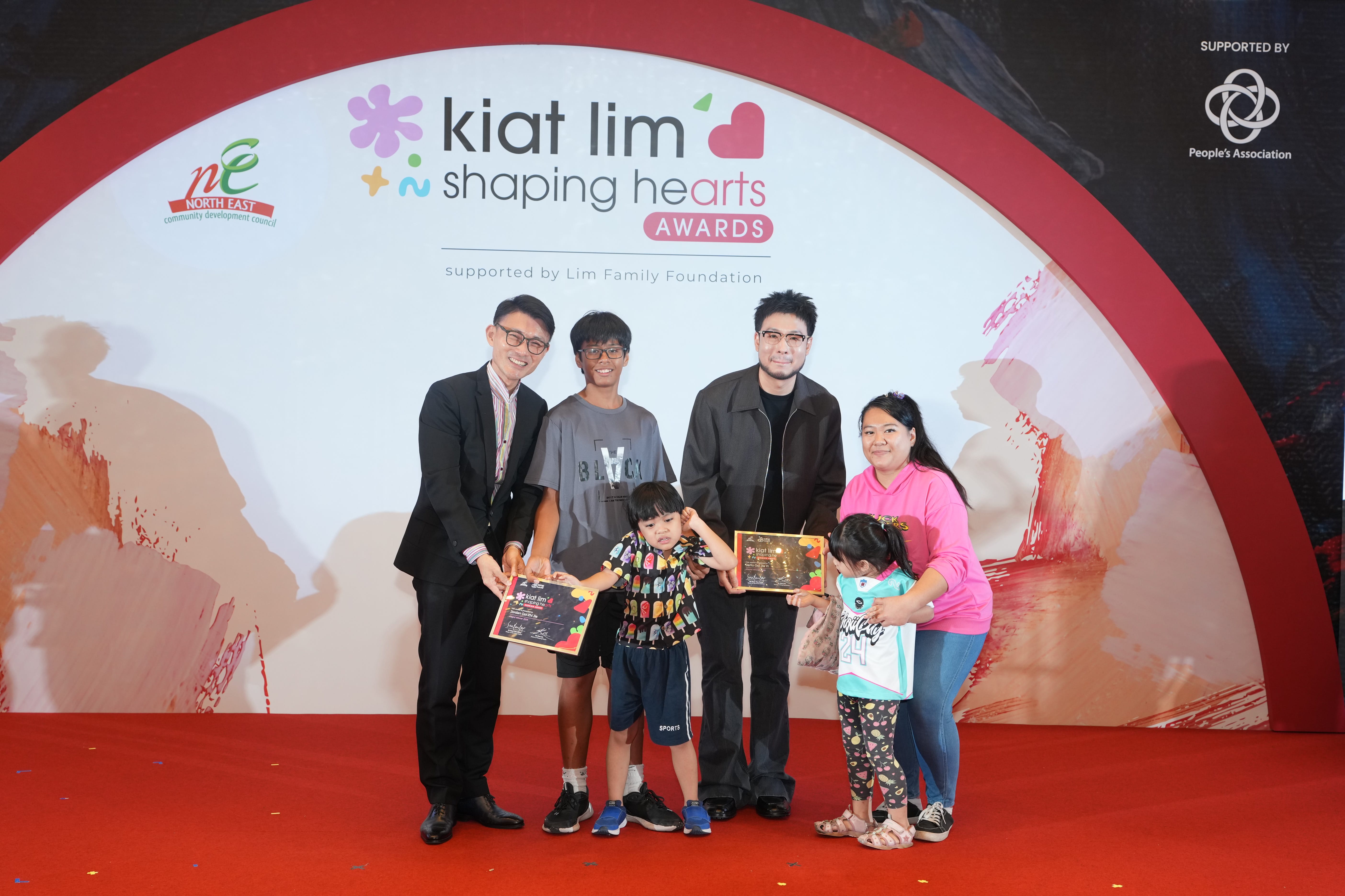 Award recipient standing on stage with the presenter, holding their certificate and posing for a group photo during the award ceremony, with a backdrop reading ‘Kiat Lim-Shaping Hearts Awards'.